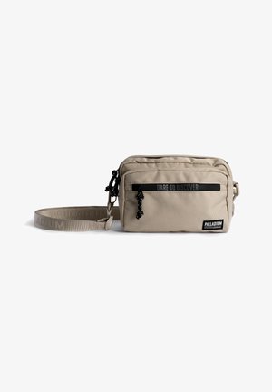 Sac bandoulière rectangulaire beige avec poche zippée sur le devant, sangle ajustable et texte "Dare to Discover" au-dessus de la poche. Étiquette de la marque "Palladium" sur le devant.