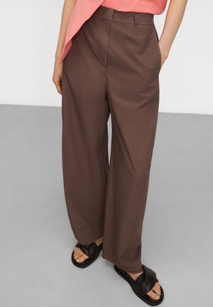 Persona che indossa pantaloni larghi marroni, top rosa e sandali neri incrociati, in piedi con le mani nelle tasche su un pavimento grigio.