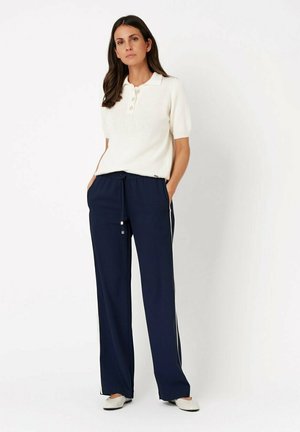 Woman wearing white short-sleeve polo shirt, navy blue wide-leg pants with side stripes, and white flats, standing against a white background.