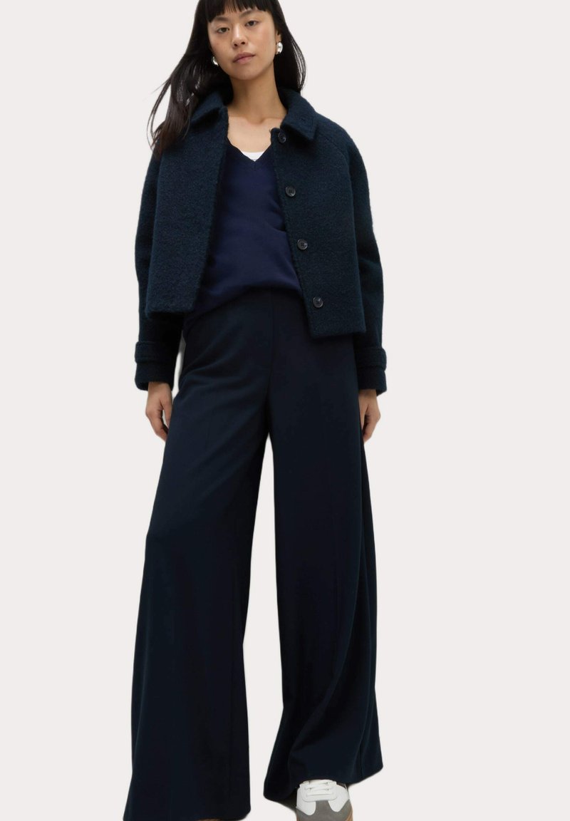 Mörkblå croppad jacka i texturerat tyg med knäppning, ihopparad med löst sittande mörkblå byxor och vita sneakers.