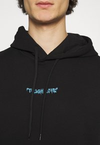 Hoodie noir avec un capuchon à cordon de serrage et du texte bleu indiquant "TOUGH LOVE" sur le devant. Tissu doux avec une coupe décontractée.