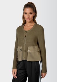 Olivgrüner Cardigan mit geripptem Textur, Knopfschluss und zwei Fronttaschen. Mit einem kontrastierenden, wildlederähnlichen Stoff am unteren Ende gestaltet.