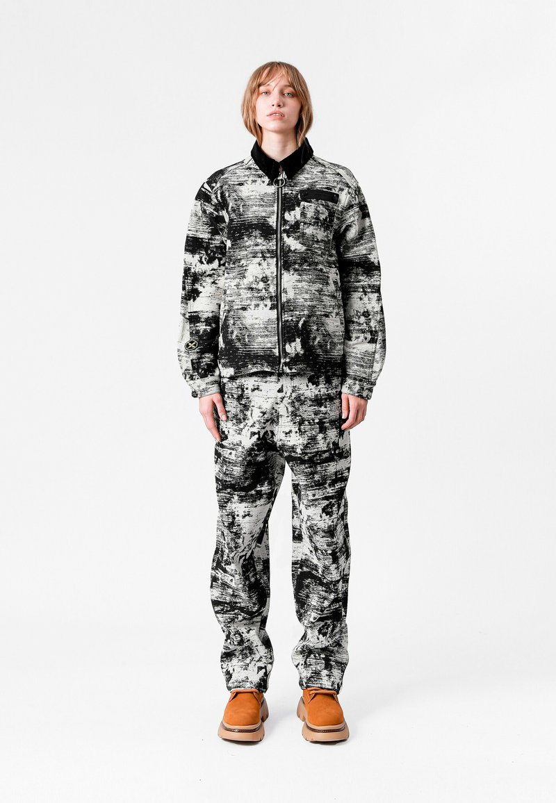 Conjunto de chaqueta y pantalones de patrón en blanco y negro, con cierre en la parte delantera, cuello y calzado marrón claro. La tela parece tener una textura con diseños abstractos.