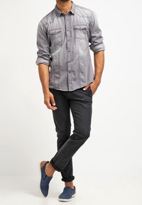 Grijze overhemd met opgerolde mouwen en twee borstzakken, gecombineerd met donkere broek en blauwe instapschoenen, wat zorgt voor een casual outfit.