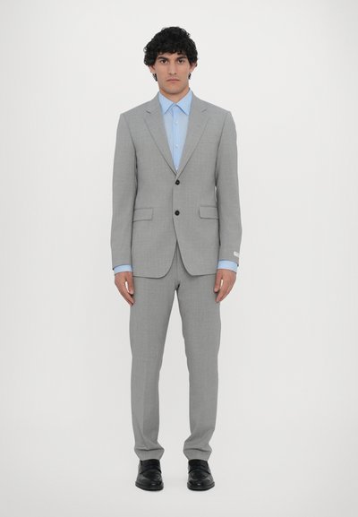 Costume gris avec une coupe ajustée, deux poches avant, boutons noirs et chemise bleue en dessous. Les chaussures sont noires, avec une texture en cuir lisse.
