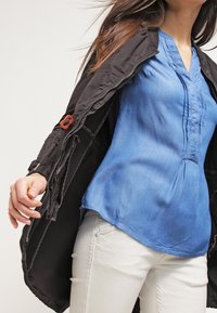 Czarna kurtka z zamkiem z przodu, rozpięta, aby pokazać lekką niebieską bluzkę z kołnierzykiem i plisowanym przodem, w zestawie z białymi spodniami.