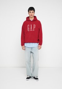 Rode capuchontrui met het "GAP"-logo, gedragen over een lichtblauw gestreept overhemd. Gecombineerd met lichtblauwe spijkerbroek en zwarte sneakers.