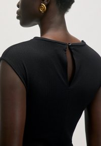 Schwarzes, geripptes Kleid mit rundem Ausschnitt, kurzen Ärmeln und einem Schlüsselloch im Rücken mit Knopfdetail. Goldene Spiralohrringe setzen Akzente.