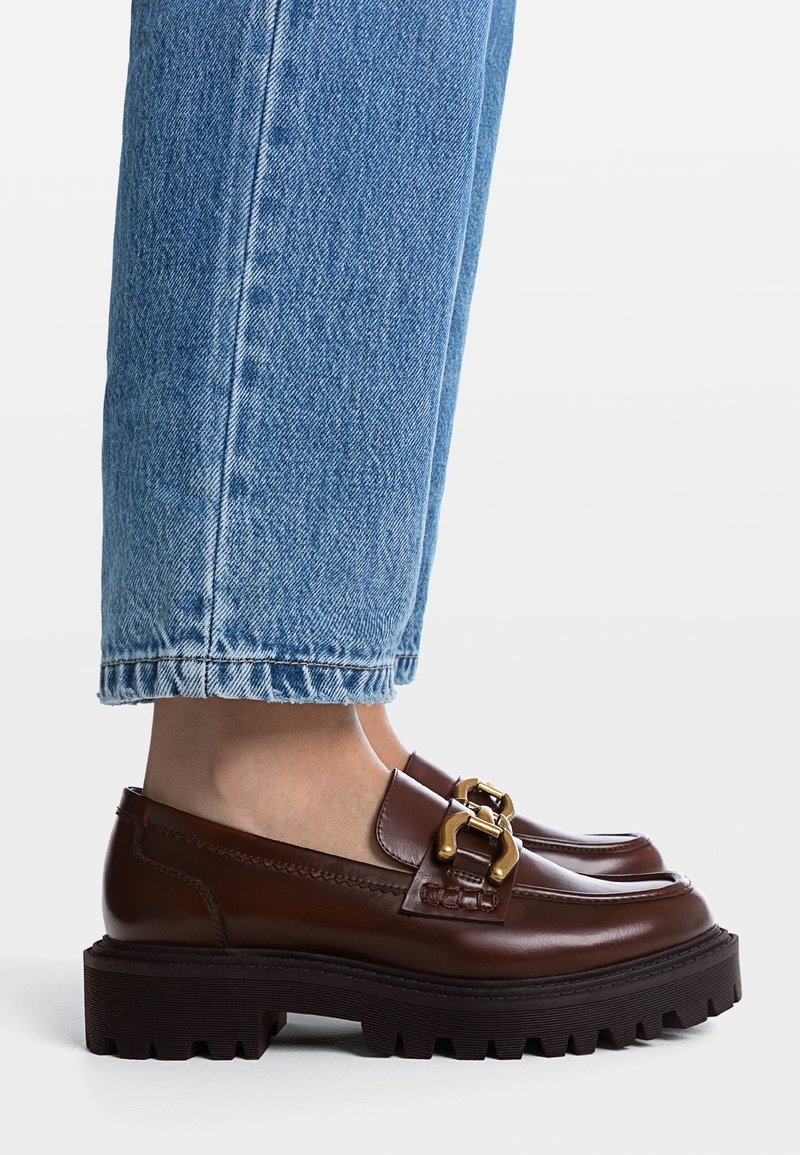 Mocassins en cuir marron avec un détail chaîne dorée, portés avec un jean bleu retroussé, sur une surface blanche plate.