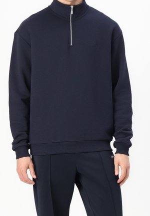 Man draagt een marineblauwe sweatvest met rits, geribbelde boorden en zoom, gecombineerd met bijpassende marineblauwe broek tegen een witte achtergrond.
