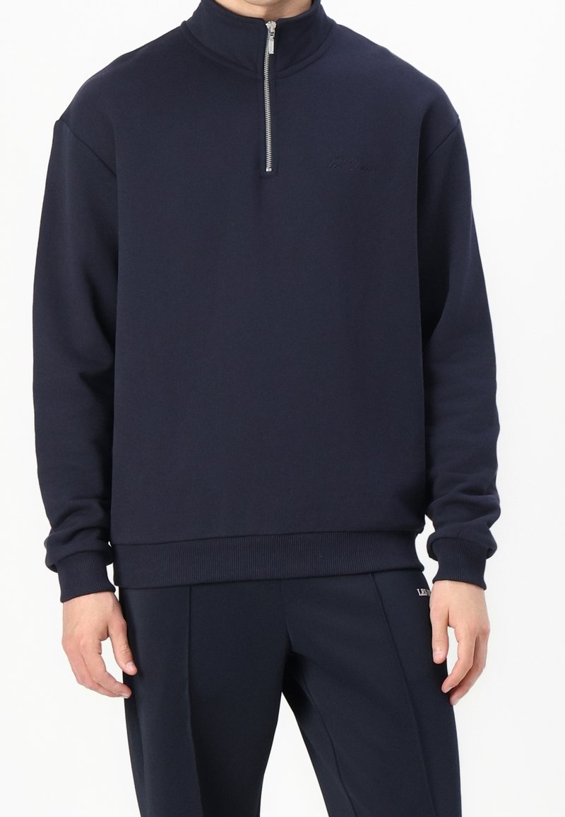 Homme portant un sweat zippé bleu marine avec poignets et bas côtelés, associé à un pantalon bleu marine assorti sur fond blanc.