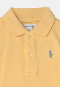 Gul polo skjorta gjord av strukturerad bomull, med en placket för två knappar och en blå broderad logotyp på vänster bröst.