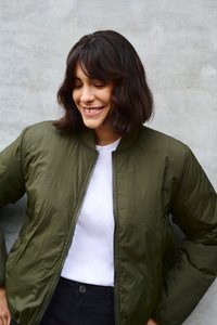 Oliivroheline bomber-jakk sileda, läikiva tekstuuriga, lukuga esiosal ja ribistatud kaelusega, valge T-särgi peal. Näha on musti pükse.