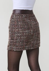 Femme portant une mini-jupe en tweed texturé taille haute avec une large ceinture marron foncé, associée à des collants noirs et un haut blanc.
