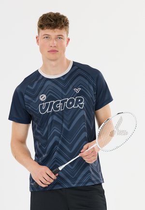 Ung mand i mørkeblå mønstret skjorte holder en hvid badmintonketcher mod en ensfarvet baggrund.