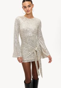 Robe courte pailletée argentée avec manches longues évasées, détail torsadé à l'avant et un léger éclat. Portée avec des bottes noires montant jusqu'aux genoux.