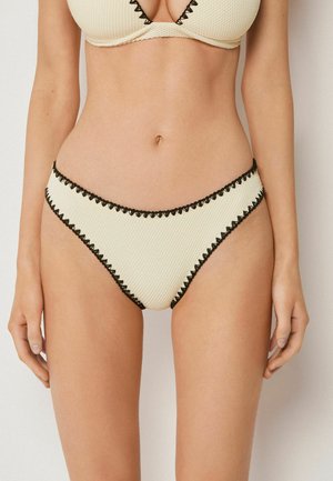 Frau trägt cremefarbenen, strukturierten Bikini mit schwarzem gewelltem Rand, gezeigt von der Brustmitte bis zum oberen Oberschenkel vor einem schlichten Hintergrund.