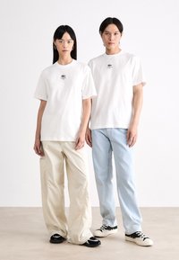Hvite bomulls t-skjorter med Lacoste-logo, kombinert med beige baggy bukser og lyseblå jeans. Skoene inkluderer svarte treningssko og beige sko.