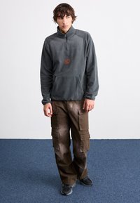 Ung man i mörkgrå fleece med kvartdragkedja, bruna cargobyxor och svarta sneakers, stående på blå matta mot vit vägg.