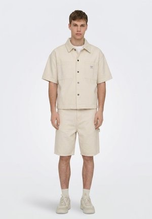 Joven de pie con camisa beige de manga corta y botones, pantalones cortos a juego hasta la rodilla, calcetines claros y zapatillas deportivas sobre un fondo blanco liso.