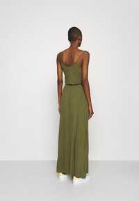 Olive green maxi dress with thin straps, a fitted bodice, and flowing skirt, paired with white shoes featuring yellow accents.