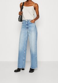 Jeans a gamba larga di un azzurro pallido abbinati a un top beige senza maniche. L'outfit include una borsa a tracolla nera e stivaletti neri con tacco quadrato.