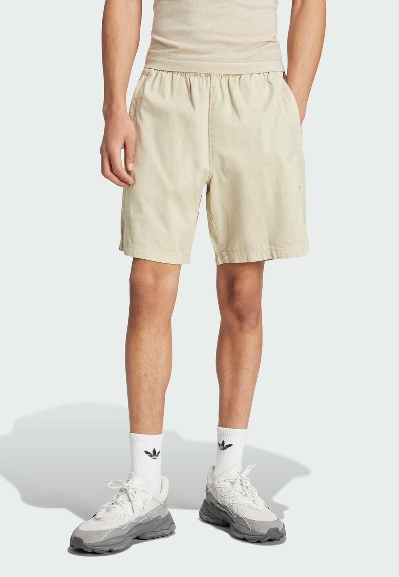 Pantaloni corti beige con cintura elastica e tasche laterali, realizzati in tessuto leggero. Indossati con sneakers bianche e calze di lunghezza media.