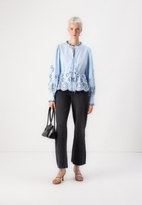 Blusa azzurra chiara con ricami floreali, vita regolabile; abbinata a jeans scuri e tacchi beige con cinturini, mentre tiene una piccola borsa nera.