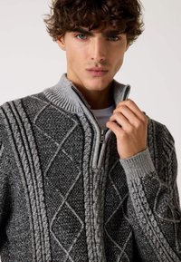 Maglione grigio lavorato a maglia con colletto alto a zip, caratterizzato da modelli testurizzati a diamante e treccia. Polsini e scollatura a costine.