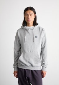 Felpa grigia chiaro in cotone con tasca a canguro frontale, cappuccio con cordino e piccolo logo scuro. Abbinata a pantaloni scuri con vestibilità ampia.