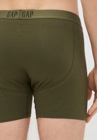 Boxeri scurți din bumbac verde, cu un bandă logo, având o croială strânsă, o textură netedă și detalii de tiv pe laturi și pe picioare.