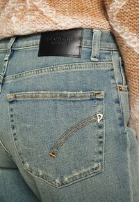 Los pantalones vaqueros de denim azul claro presentan una etiqueta de cuero negro, un diseño de bolsillo único con un detalle metálico, y costuras visibles en la parte trasera.
