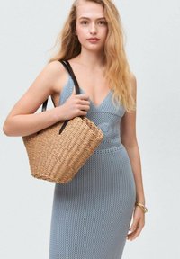 Blå stickad klänning med V-ringning och figurnära passform. Modellen håller en beige vävd handväska med svarta handtag. Slät textur med ett ribbat mönster.