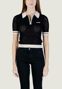 Femme portant un haut noir en maille à manches courtes avec col et poignets blancs, fermeture éclair à l'avant, de la marque "GUESS", associé à un pantalon noir taille haute.