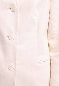 Blazer de color beige claro con un patrón texturizado, cinco botones en fila y un bolsillo en el pecho con borde piped.