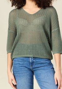 Pull over en maille lâche vert olive avec un col en V et des manches 3/4, présentant un design texturé en tricot à mailles ouvertes. Associé à un jean en denim bleu.