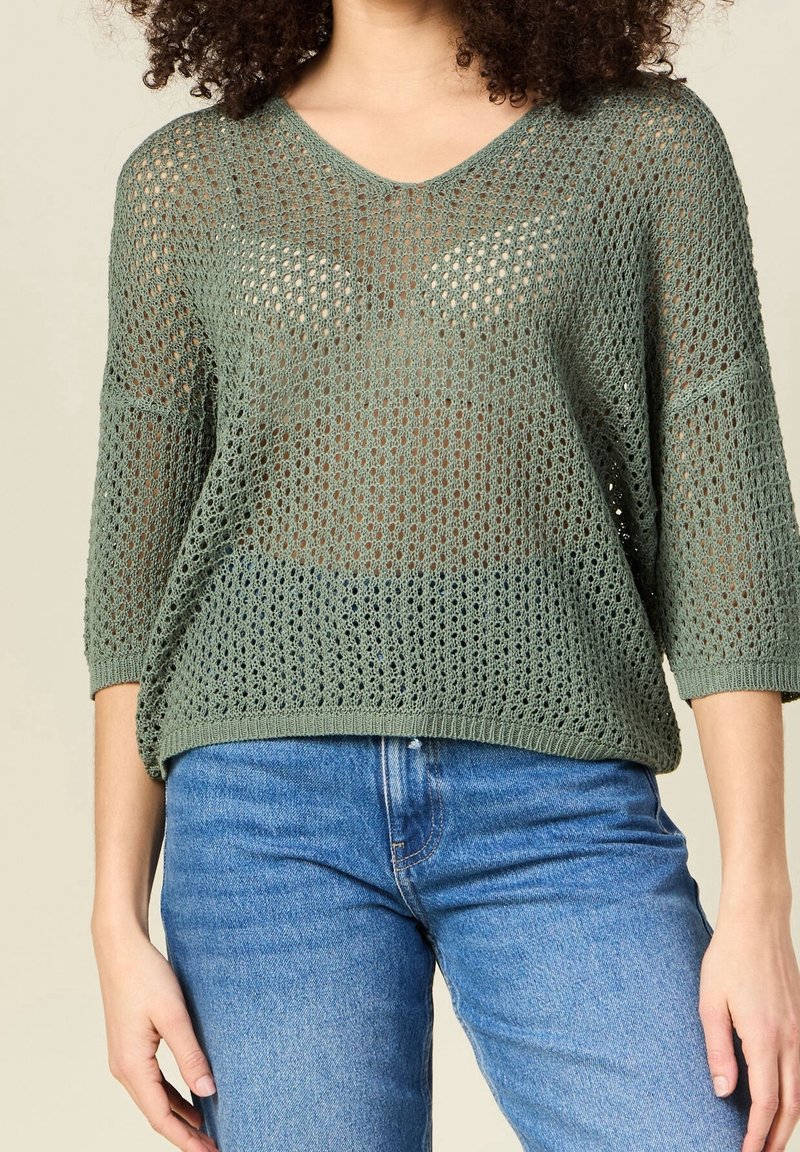 Pull over en maille lâche vert olive avec un col en V et des manches 3/4, présentant un design texturé en tricot à mailles ouvertes. Associé à un jean en denim bleu.