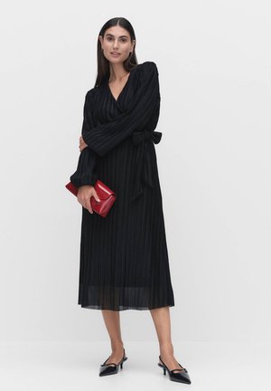 Zwarte, lange mouw wikkeljurk met verticale strepen, bijpassende stoffen riem en een hem van midi-lengte. Accessoires zijn een rode clutch en zwarte hakken.