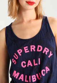 Mörkblå tanktop i lätt tyg med palmträdsmönster, med fet rosa text: "SUPERDRY CALI MALIBUCA" över framsidan.