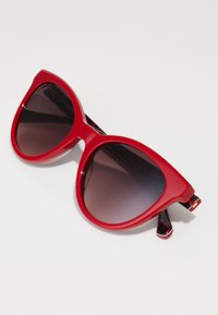 Red cat-eye sunglasses with dark gradient lenses; glossy plastic frame; patterned black and red temples.