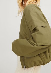 Kvinde med blond hår iført en oversized olivengrøn bomberjakke over en lys strikket kjole, der står mod en ensfarvet lys baggrund.