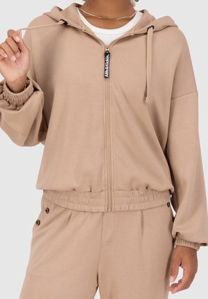 Femme portant un sweat à capuche beige zippé et un pantalon assorti avec des poignets et une ceinture élastiques, tenant le cordon de la capuche de la main gauche.