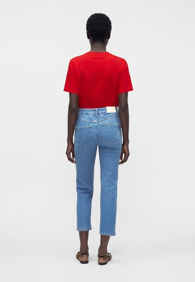 Personne debout, de dos, portant un t-shirt rouge à manches courtes, un jean court bleu clair et des sandales imprimé léopard sur fond blanc.