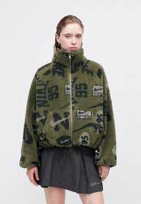 Olive rohelises fleece-jakk, millel on kõrge krae, tõmblukk, eesmine tasku ja mustad graafilised mustrid. Tekstuuriga ja ülemõõduline disain.