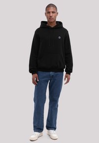Black hoodie with a front pocket, drawstrings, and a small logo. Paired with blue jeans and white sneakers, featuring a relaxed fit.