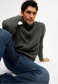 Grå ribbad zip-up tröja med hög krage, matchad med blå denimjeans. Modellen sitter i en avslappnad pose mot en neutral bakgrund.