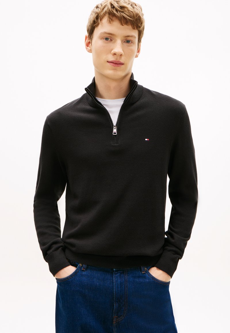Jeune homme aux cheveux châtains clairs portant un pull noir à fermeture éclair sur une chemise blanche et un jean bleu, debout les mains dans les poches.