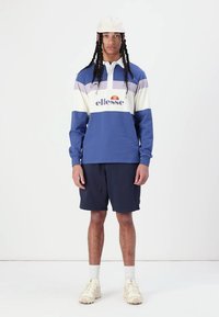 Camisa de polo de mangas longas azul, branca e lavanda com logotipo e riscas, combinada com calções azul-marinho e ténis bege numa pessoa.