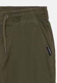 Pantalons en coton vert olive avec taille élastique, cordon de serrage, poches latérales et petite étiquette noire de la marque Quiksilver près de la poche.