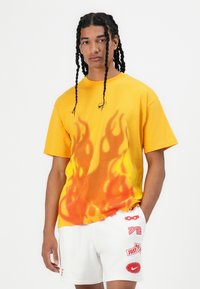 Tricou galben cu un design de flacără care se estompează în portocaliu. Prezintă un logo mic pe piept. Asortat cu pantaloni scurți albi decorați cu diverse grafice.